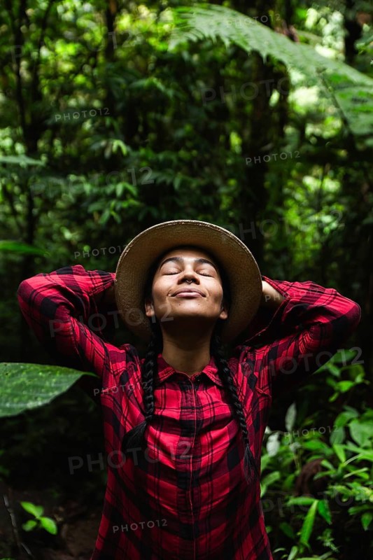 An Ecuadorian woman enjoys the natural beauty of the Choco Andino rainforest in Ecuador. Wearing a red plaid shirt and a hat, she relaxes among lush greenery, breathing in the fresh air of this biodiverse region with eyes closed