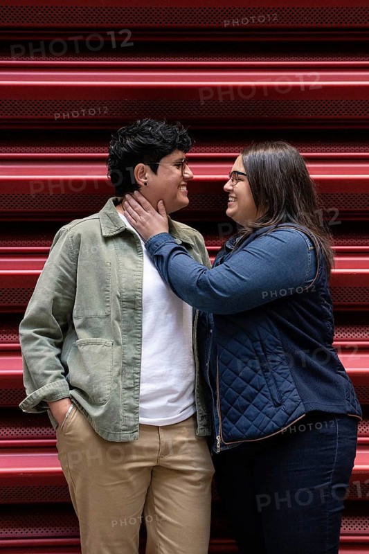 A joyous moment captured between a curvy lesbian couple standing closely outdoors. They gaze lovingly at each other, exuding happiness and affection against a vibrant red background