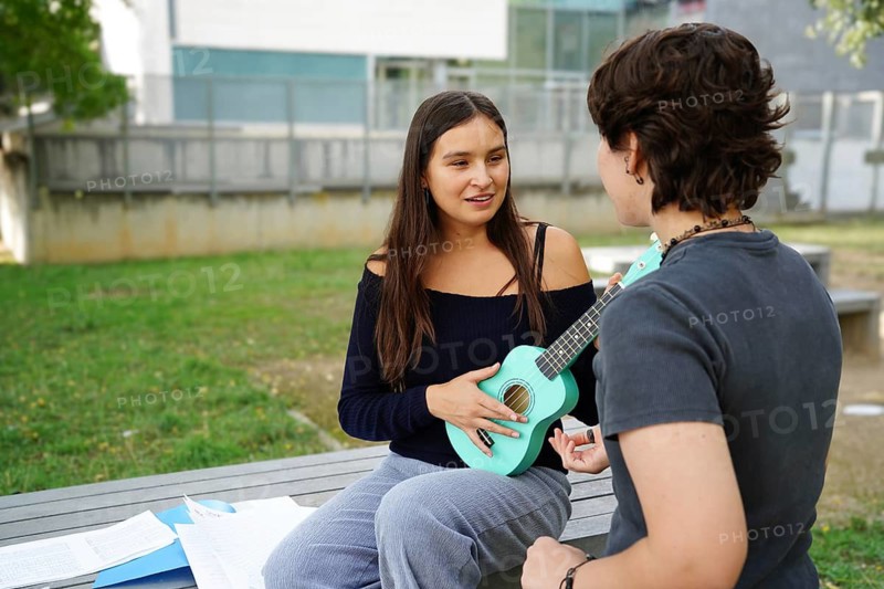 Two teenagers, a non-binary person and a female, engage in a vibrant conversation while sitting on a bench at school. The woman holds a turquoise ukulele, contributing to an inspiring musical exchange
