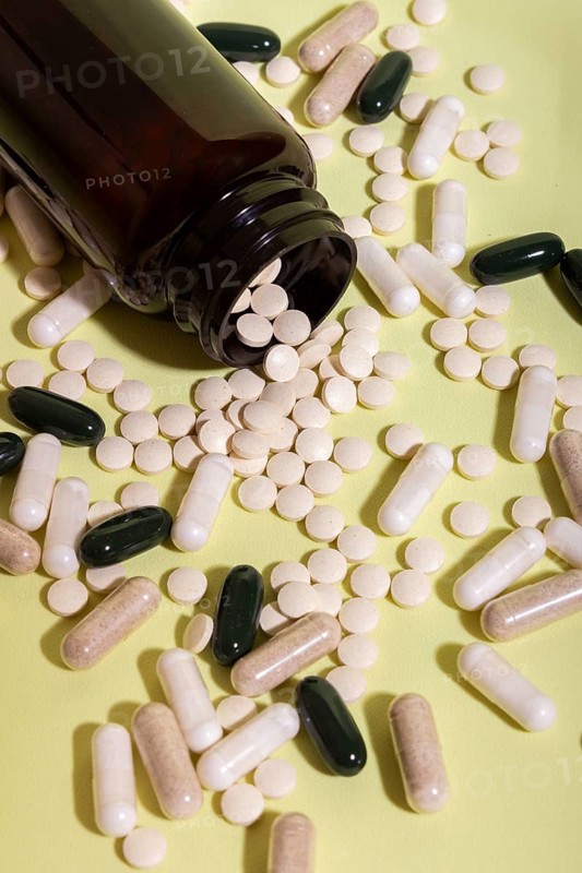 An open glass bottle spills assorted pills and capsules onto a light green surface. The variety of shapes and colors represents diverse medicinal and sanitary materials