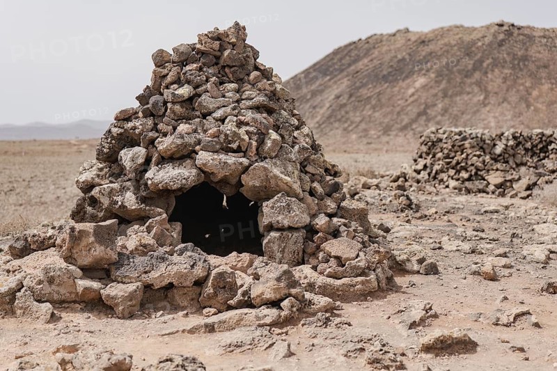 Traditional stone hut amidst a dry, barren landscape in Fuerteventura, exemplifying the arid textures and natural formations typical of the Canary Islands' summer landscapes