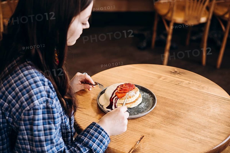 A woman in a plaid shirt sits at a wooden table, enjoying a plate of fluffy japanese pancakes topped with fresh strawberries and syrup. The cozy cafe setting adds warmth