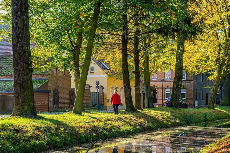Woman in red jacket walking through a park with autumn coloured trees at the river Nieplitz, behind her old brick houses, Treuenbrietzen, Fläming, Brandenburg, Germany