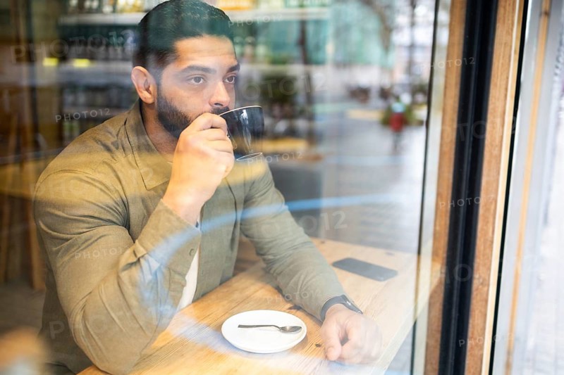 A man sits at a wooden table by a cafe window of the city, sipping coffee. The ambiance is serene, with reflections in the glass. The setting suggests contemplation and relaxation