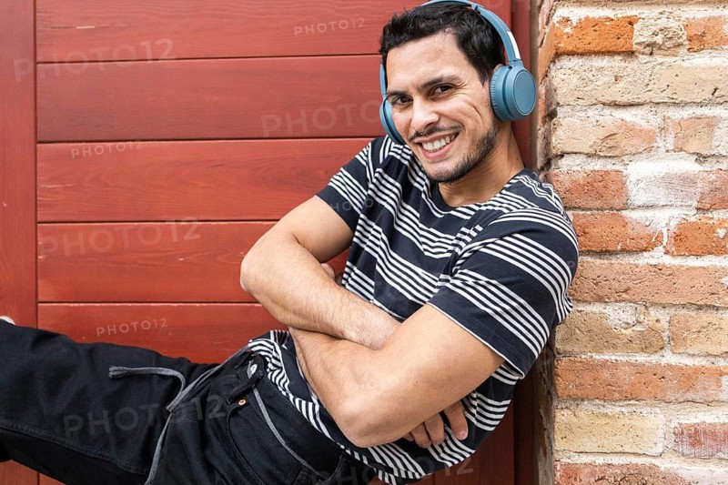 A cheerful cuban man listens to music with blue headphones, leaning against a brick wall. He is casually dressed in a striped shirt, exuding joy and relaxation