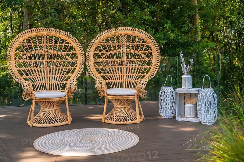 Two wicker peacock chairs and white decorations stand on a wooden deck, creating a bohemian atmosphere for a wedding ceremony