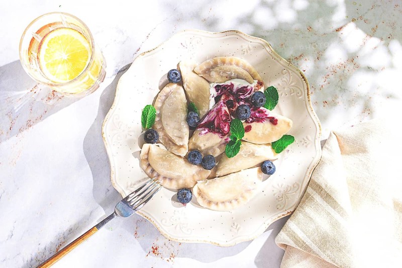 Handmade dumplings filled with sweet ingredients, arranged beautifully on a white plate, topped with berry sauce, fresh blueberries, and mint leaves, ready to enjoy