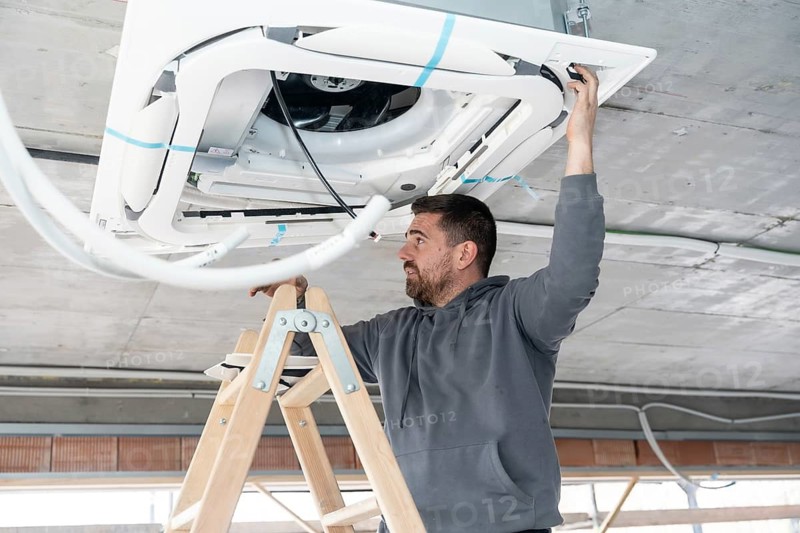Professional hvac technician installing a ceiling mounted air conditioning unit on a ladder, working to integrate the system into a building under construction