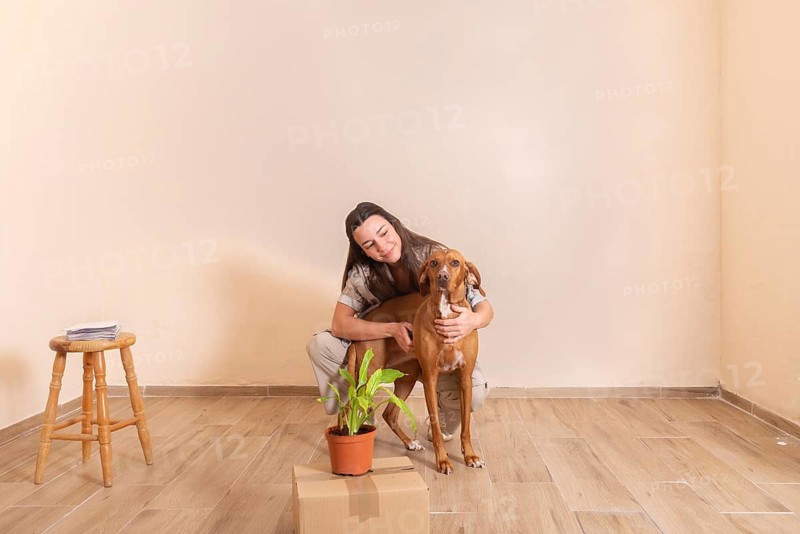 A young woman lovingly embraces her dog in a sparsely furnished room, surrounded by a stool, a potted plant, and a cardboard box, conveying warmth and companionship