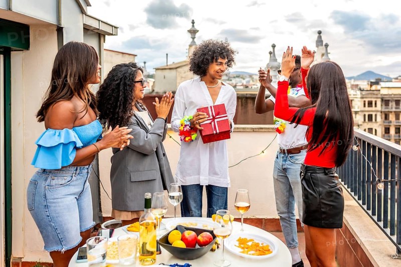 Multi-ethnic young chic friends applauding while man receiving a birthday gift during rooftop party during sunset