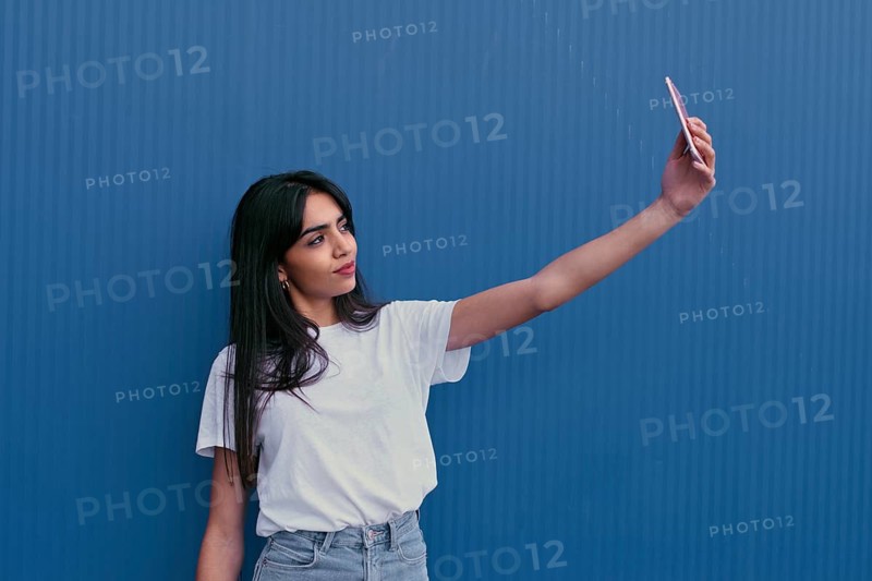 A young woman in a white shirt takes a selfie against a vibrant blue wall. Her confident expression captures the essence of modern self expression and digital culture