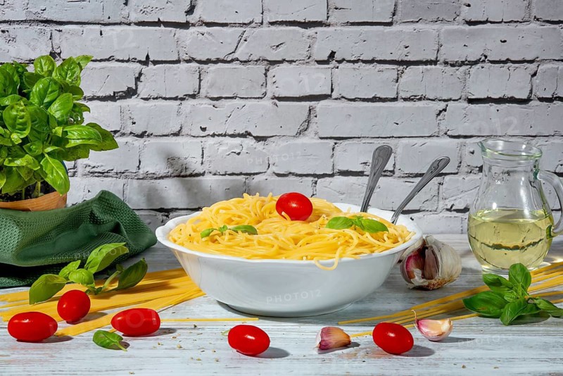 Bowl of cooked spaghetti surrounded by tomatoes, basil, olive oil and garlic on a table