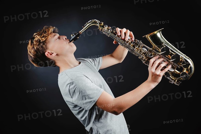 A teenage boy passionately plays the saxophone in a studio setting with a dark, black background, focusing intently on his musical performance