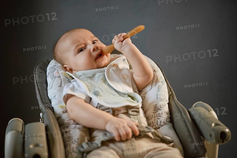 A baby sits in a high chair with a bib on and holds a wooden spoon. The baby is focused on the spoon while enjoying mealtime at home. The background is simple and neutral in color