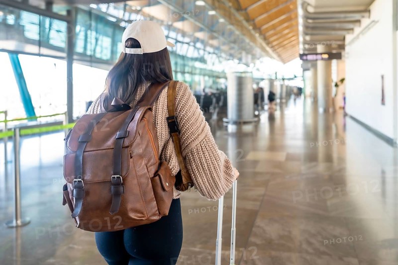 Woman walking from behind wearing a cap and a knit sweater, carrying a brown backpack and pulling a suitcase inside a bright airport terminal building