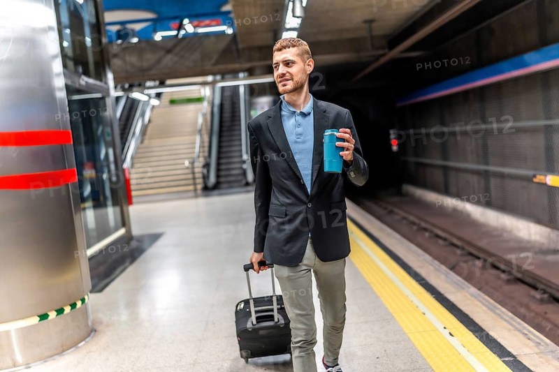 Young businessman walking through a bustling subway station, carrying a suitcase and sipping coffee during his morning commute