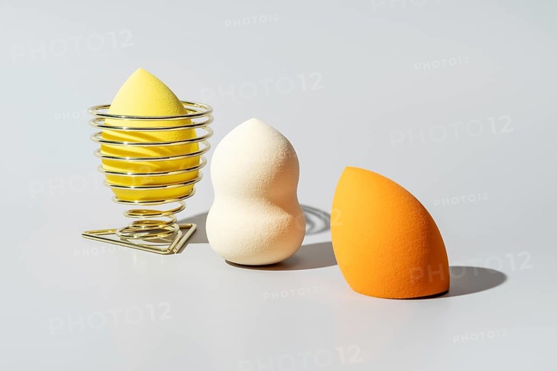 A collection of cosmetic sponges in diverse shapes and vibrant colors, including yellow in a holder, white, and orange, arranged on a light surface