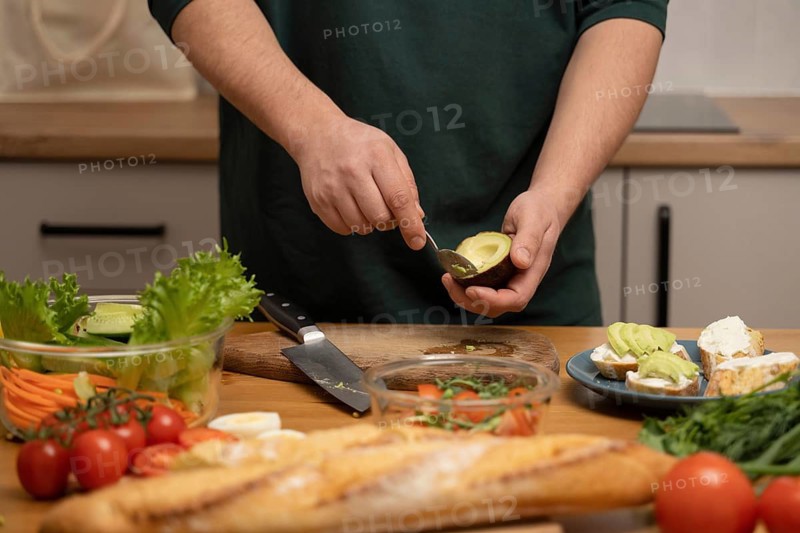 A person skillfully prepares an avocado, surrounded by fresh vegetables and bread on a kitchen counter. A delicious spread is ready for a healthy meal