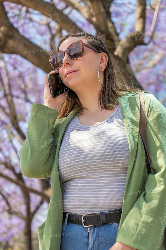 Low-angle vertical shot of a happy woman in a green jacket, talking on the phone beneath blooming jacaranda trees in an urban setting. The bright purple flowers and sunlight enhance the spring atmosphere
