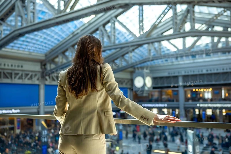 Young businesswoman with long brown hair, wearing a beige suit, standing on a balcony, observing commuters and modern architecture inside a large, bustling train station in new york city