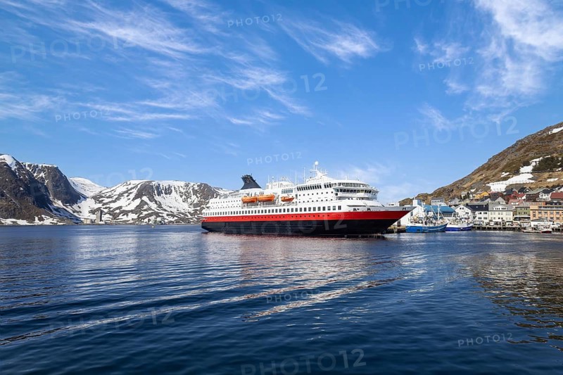 A large Norwegian Cruise ship with red hull docked at Honningsvag port, Norway, with snow-covered mountains and a small Arctic town visible under a bright blue sky