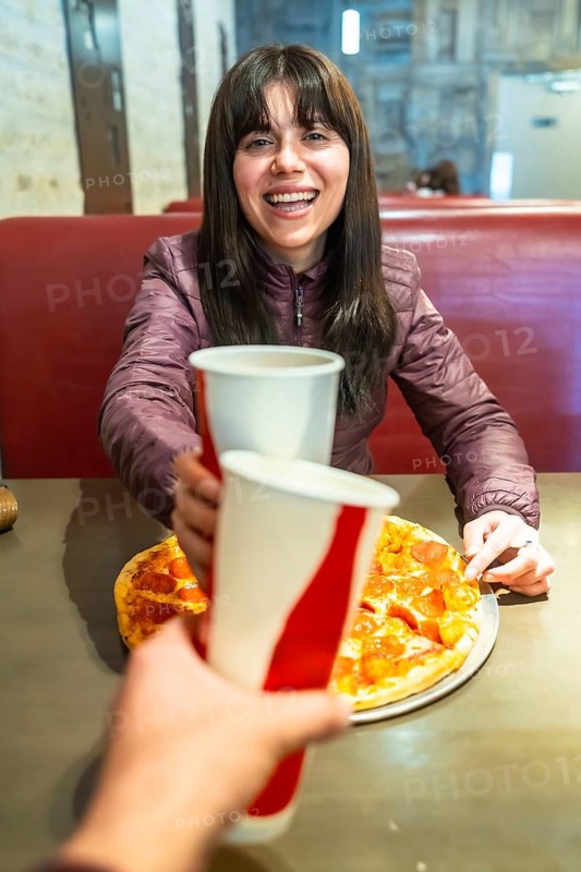 Smiling woman enjoying pizza at a casual restaurant, clinking paper cups with a friend in a candid moment of friendship, food, and relaxed weekend fun