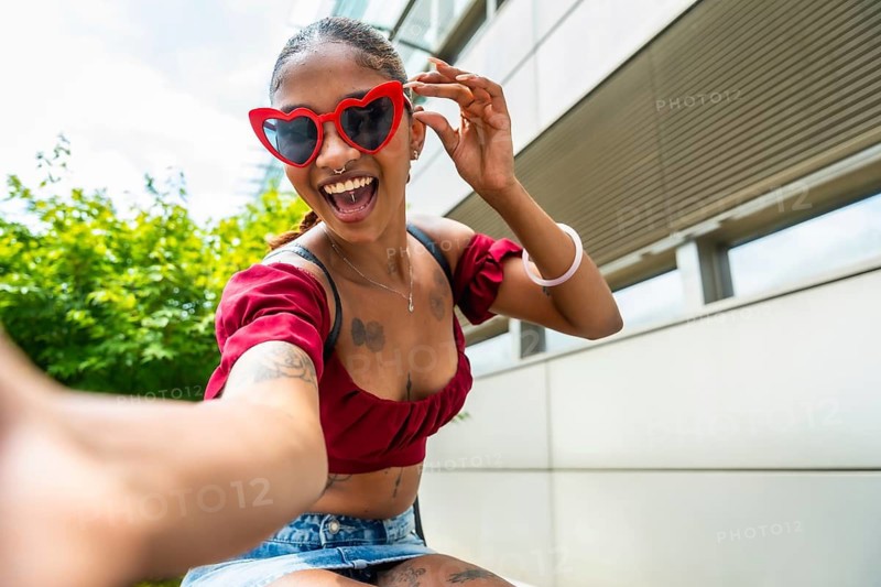 Young woman with heart shaped sunglasses and a wide smile enjoying a moment of self expression and fun, capturing a happy selfie outdoors