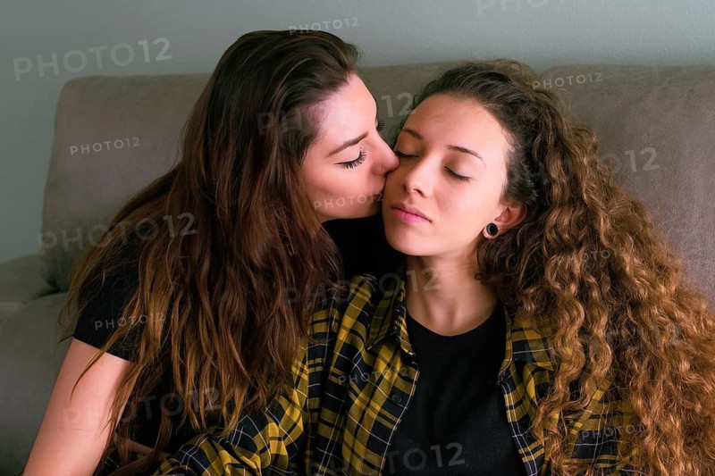 A female gay couple shares a loving embrace on a couch. The woman on the left kisses the forehead of the other, who has her eyes closed and is smiling gently