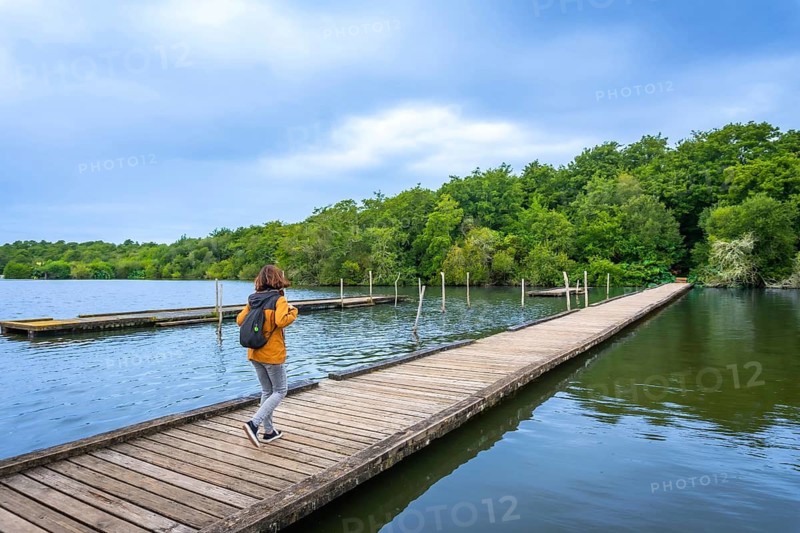 Woman with backpack walking along a wooden pier on lake azur in landes, france, enjoying a peaceful outdoor summer stroll amid blue skies, water and green forest scenery
