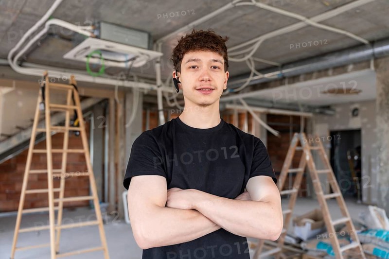 Young man working as an hvac technician standing confidently with crossed arms, smiling at a construction site with exposed wires and air conditioning units
