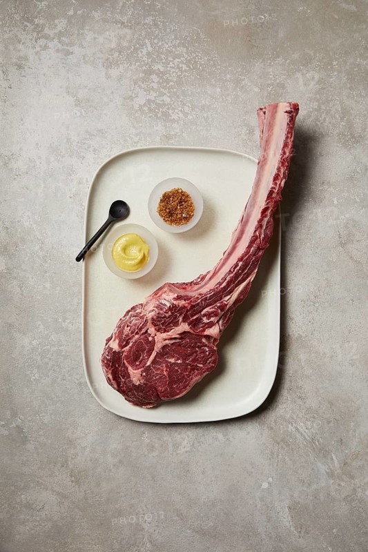 Top-down view of a raw tomahawk steak presented on a ceramic tray, accompanied by a small bowl of mustard, spices, and a black spoon. A premium cut ready for gourmet cooking