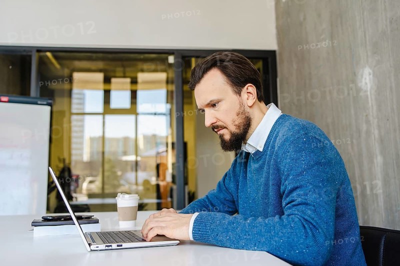 A focused oman works on a laptop in a modern office setting, surrounded by glass walls and a coffee cup nearby, highlighting productivity in a contemporary workspace