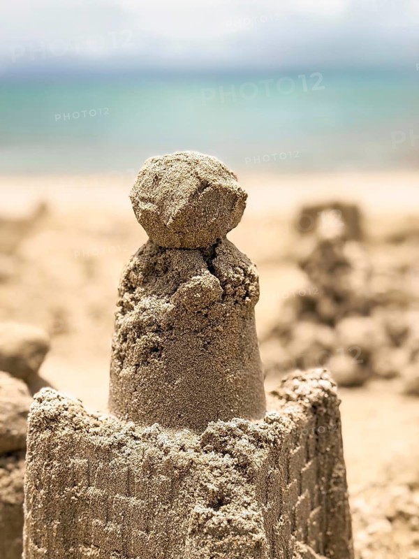 A detailed sand castle stands proudly on the seaside, with intricate designs and a smooth dome, against a backdrop of clear blue skies and distant ocean waves