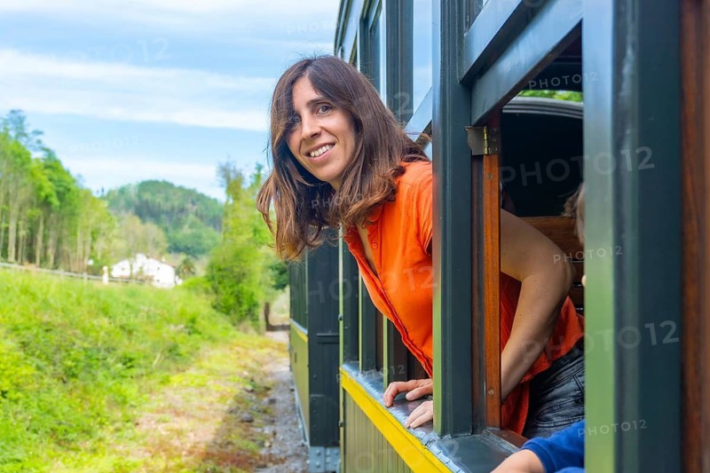 A woman is sitting in a train car with her head out the window. She is smiling and looking out at the scenery