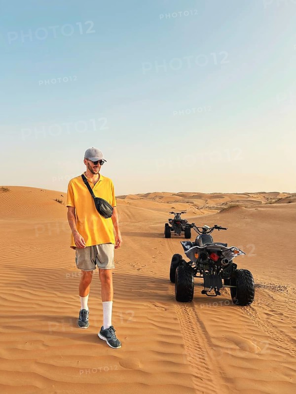 A happy smiling man in a yellow shirt and cap walks near an all terrain vehicle in the expansive Wahiba Sands Desert, Oman, showcasing adventure and exploration in a barren yet beautiful landscape