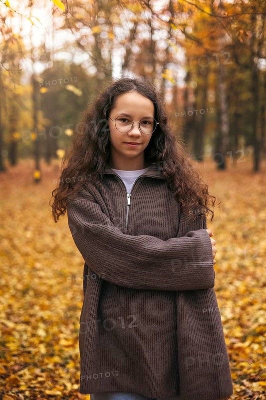 A girl in a cozy brown sweater stands amid a forest filled with vibrant autumn leaves. Her serene expression captures the essence of the fall season