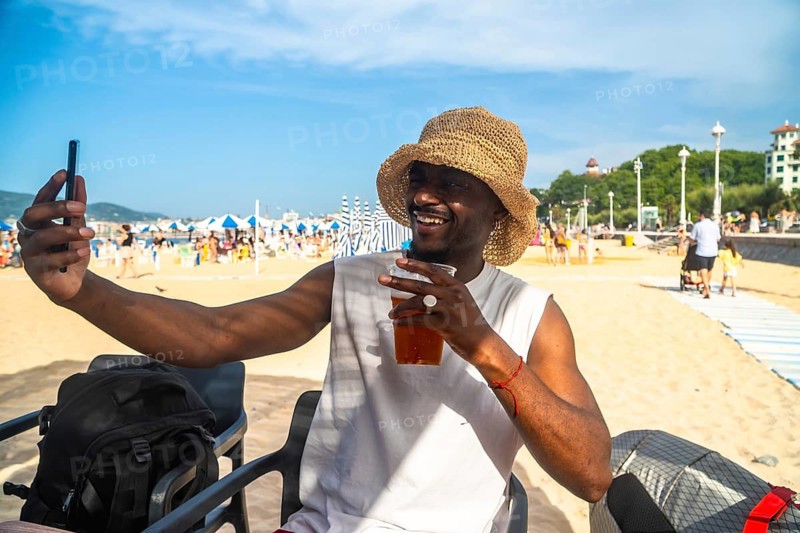 Smiling young man enjoying a relaxing summer vacation, holding a refreshing drink and taking a selfie on a vibrant beach with people and parasols in the background