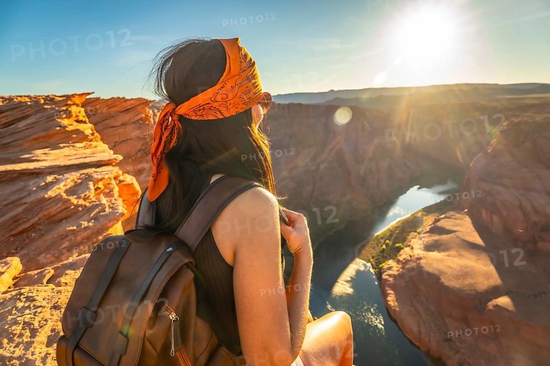 Woman traveler with bandana and backpack sitting on a cliff edge, carefully looking at the scenic colorado river winding through the sandstone canyon during golden hour