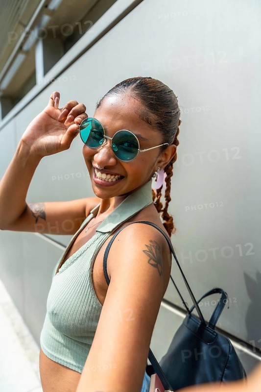 Smiling young woman adjusting her round blue sunglasses, wearing a light green crop top and braided hair, standing outside a modern building and expressing joy