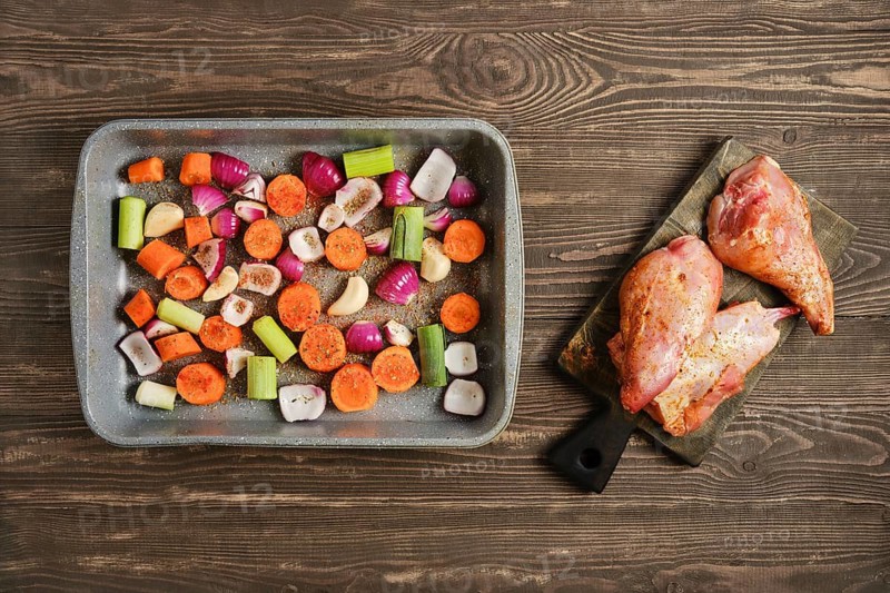 A tray holds chopped vegetables including carrots, celery, garlic and onions along with seasoned rabbit meat on a cutting board. This scene is set on a wooden table in a kitchen during daytime