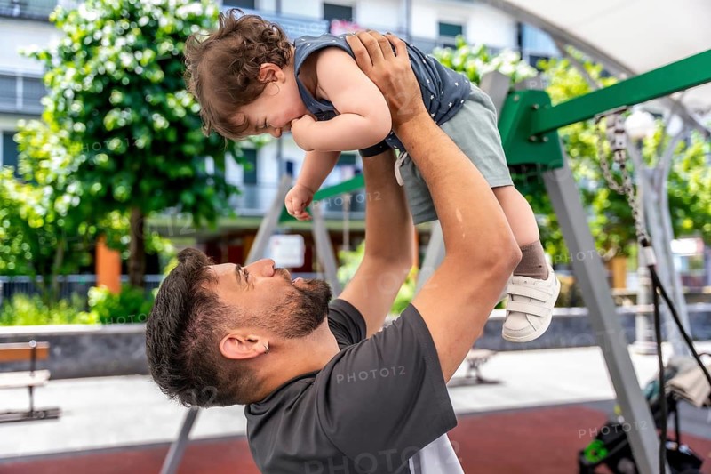 Father lifting up his young son, creating a joyful moment of play and bonding in an urban park playground on a sunny day, representing family connection and happiness