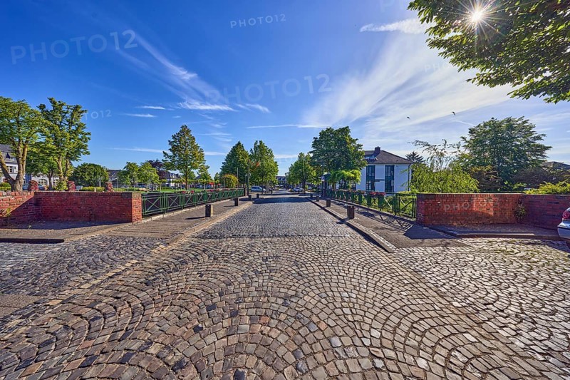 Car bridge and footbridge, general buildings, houses, metal railings, barrier bollards, sidewalk, roadway, road, paving stones, trees, sun backlight, blue sky, cirrus clouds, cirrostratus clouds, Feldstraße, Friedensplatz, Wachtendonk, Kleve district, North Rhine-Westphalia, Germany