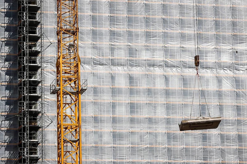 Essen, North Rhine-Westphalia, Germany - Construction crane lifts concrete section in front of scaffolded office building site facade