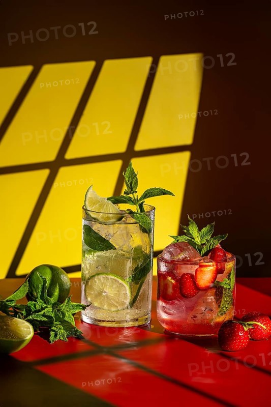 Refreshing mojito cocktails displayed on a vibrant table. A classic mojito with lime and mint alongside a strawberry mojito with fresh strawberries. Bright yellow and red background enhances the scene