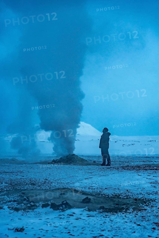 A man stands in the snow looking at a large plume of smoke. The sky is a deep blue color, and the snow is white. The scene is quiet and peaceful, with the man being the only person in the image