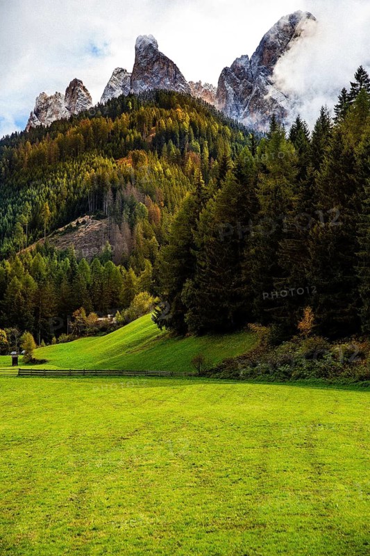 Mountain landscape with green fields and thick forests, under cloudy skies and autumn vegetation, view of the Geisler Group mountain from the Funes valley. Funes, Trentino Alto Adige, Italy
