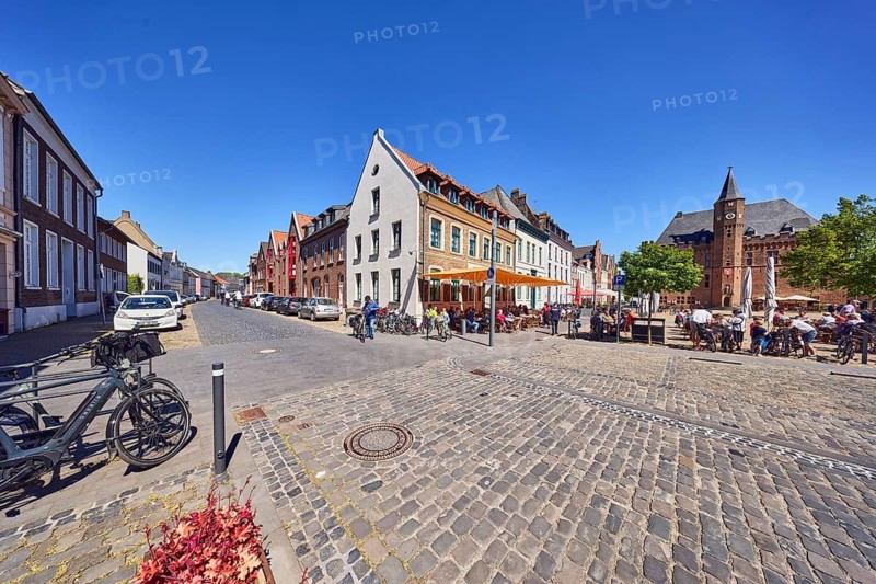 Old town, historic city center, historic residential buildings and commercial buildings, restaurant, outdoor area, town hall, square, street, paving stones, parking strips with cars, bicycles, trees, gastronomic visitors, cyclists and pedestrians as a secondary motif, blue sky, cloudless, Kesselstraße confluence with Markt, Kalkar, Kleve district, North Rhine-Westphalia, Germany