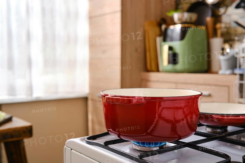 A red cast iron pot with enamelled coating on a gas stove in a kitchen. The kitchen has a blend of wood and light colors. Natural light comes through a window, creating a warm atmosphere for cooking