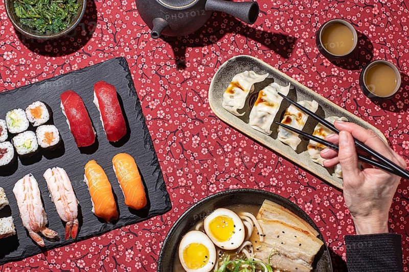 Artistic composition of a traditional Japanese feast, including sushi, dumplings, and ramen, with a visible hand using chopsticks, creating a natural and engaging atmosphere