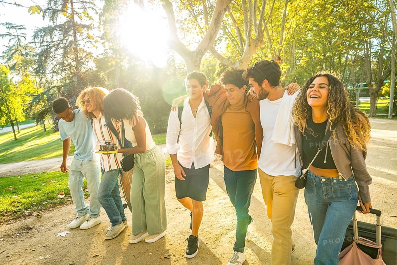 Multiracial young students walking together embracing from the back along a sandy path at park